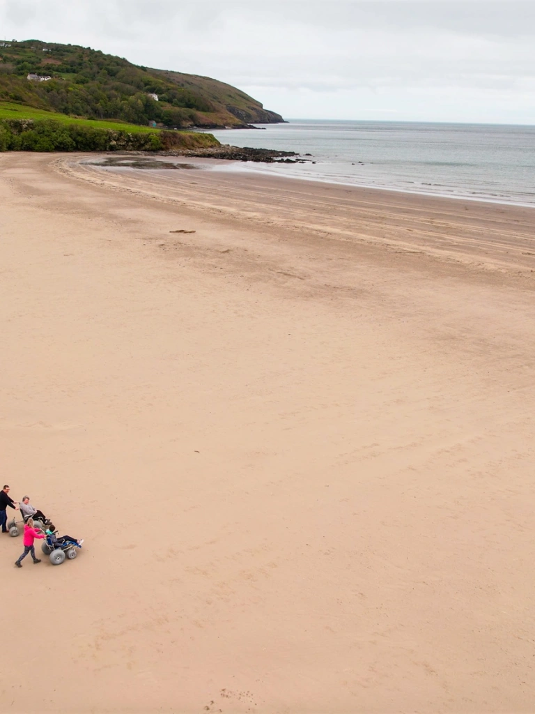 A drone shot of a wide sandy beach with two people using beach wheelchairs.