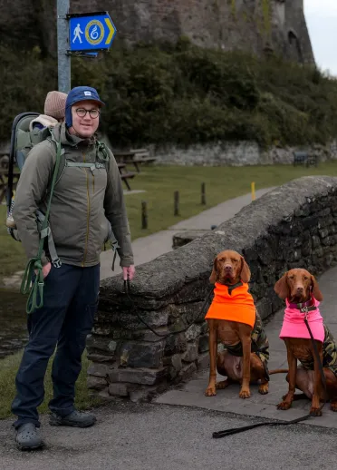 Man with baby carried on his back and two Vizslas dogs on bridge with Laugharne Castle in background.
