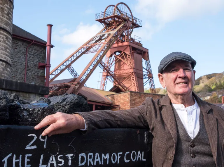 A former miner smiling at the camera with a tower colliery behind him.