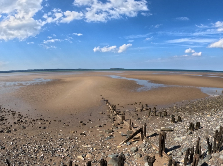 View across a sandy beach out to sea with the tide out and a blue sky above.