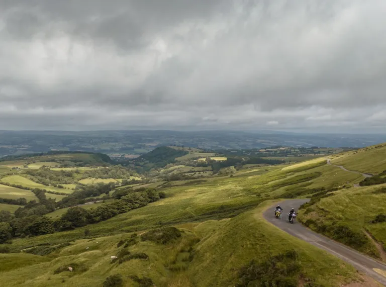 two motorbikes on road with surrounding countryside on cloudy day.