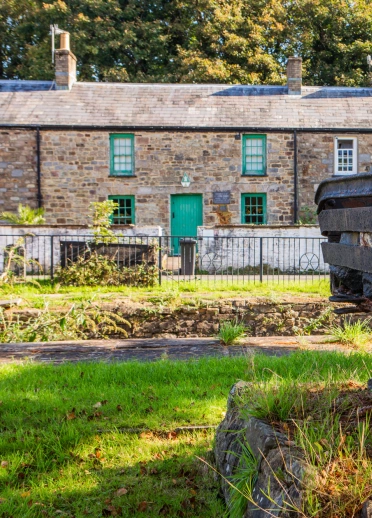 A sunny exterior shot of the row of stone cottages with a dark iron and wood coal dram in the foreground.