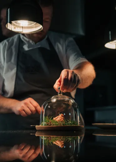 A male chef putting a small glass dome over a plate of food.