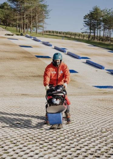 A child in an adapted sled / ski-pram being pushed by a man on a dry ski-slope.