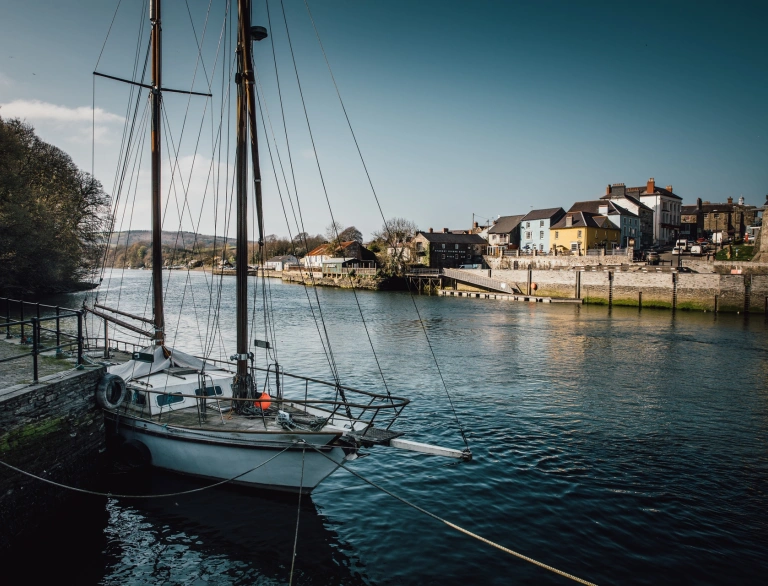 A wide river with a sailing boat in the foreground and a quay in the background.