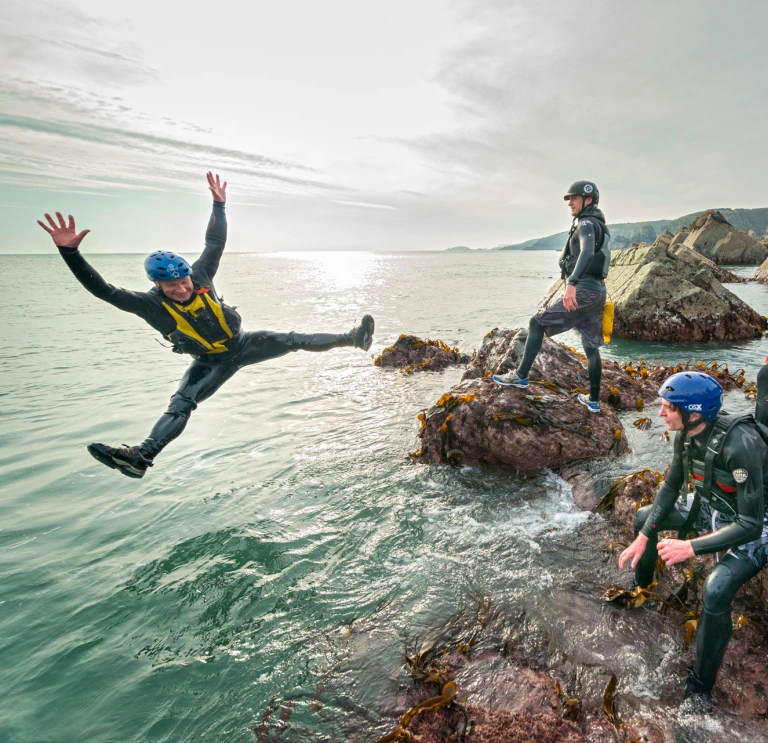 Group coasteering on rocks with one member star jumping into the sea.