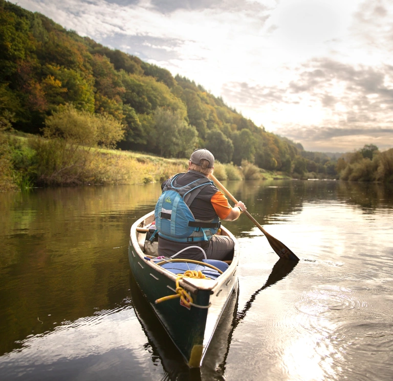 A man using a canoe on a wide, calm, tree-lined river.