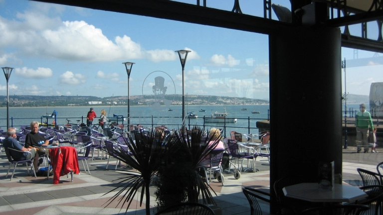 Outside seating area with sea in background, viewed through cafe window.