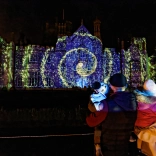 man and woman holding children watch light show projected onto manor house.