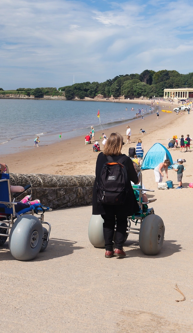 Three females pushing all-terrain beach wheelchairs down slope towards sandy beach.